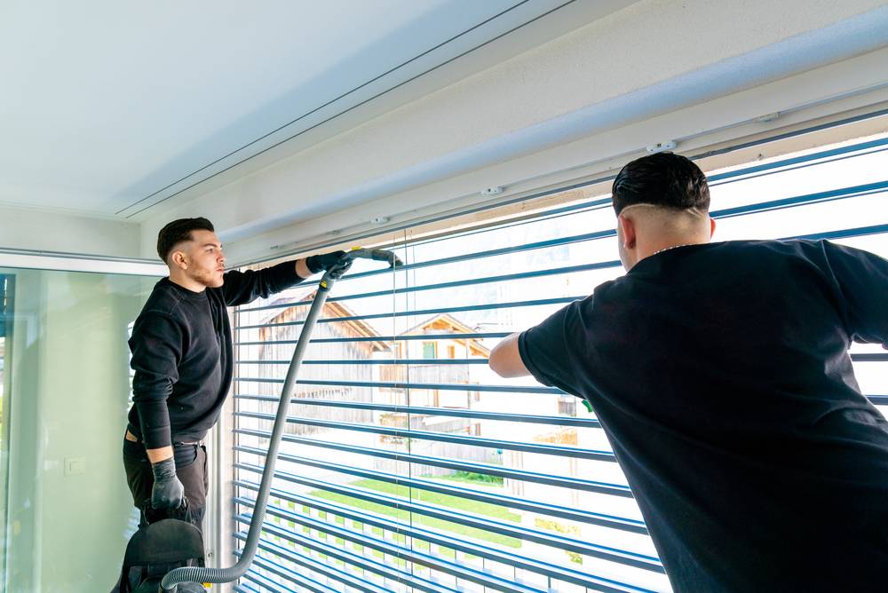 Zwei GDM-Mitarbeiter reinigen Fensterfront und Jalousien in einem hellen Raum